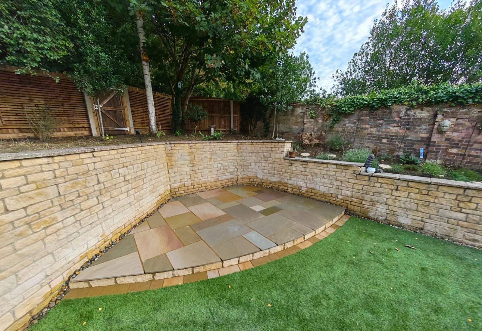 Multi-tone Indian sandstone patio with a curved raised edge in the corner of a walled garden in Weldon